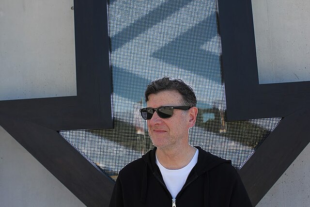 Volition label founder Andrew Penhallow stands in front of a window shaped like an arrow pointing downward. He wears stylish sunglasses, a white tee shirt and black hoodie.