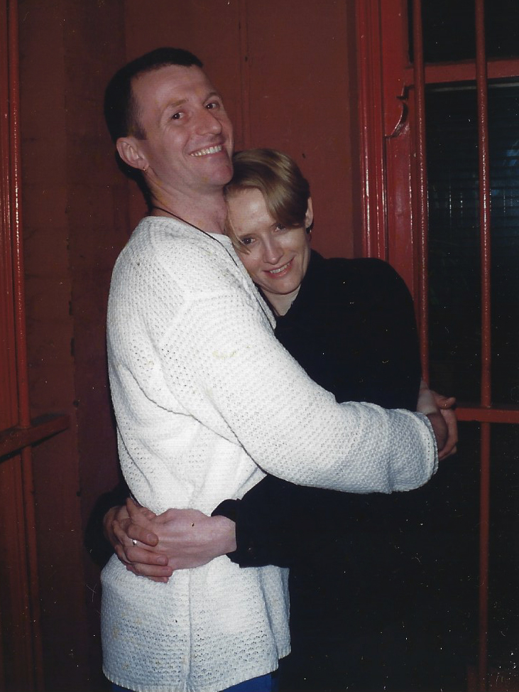 The image shows the late Robert Racic on the left in a white sweater hugging Kathy Naunton on the right. Kathy is wearing a black sweater. 