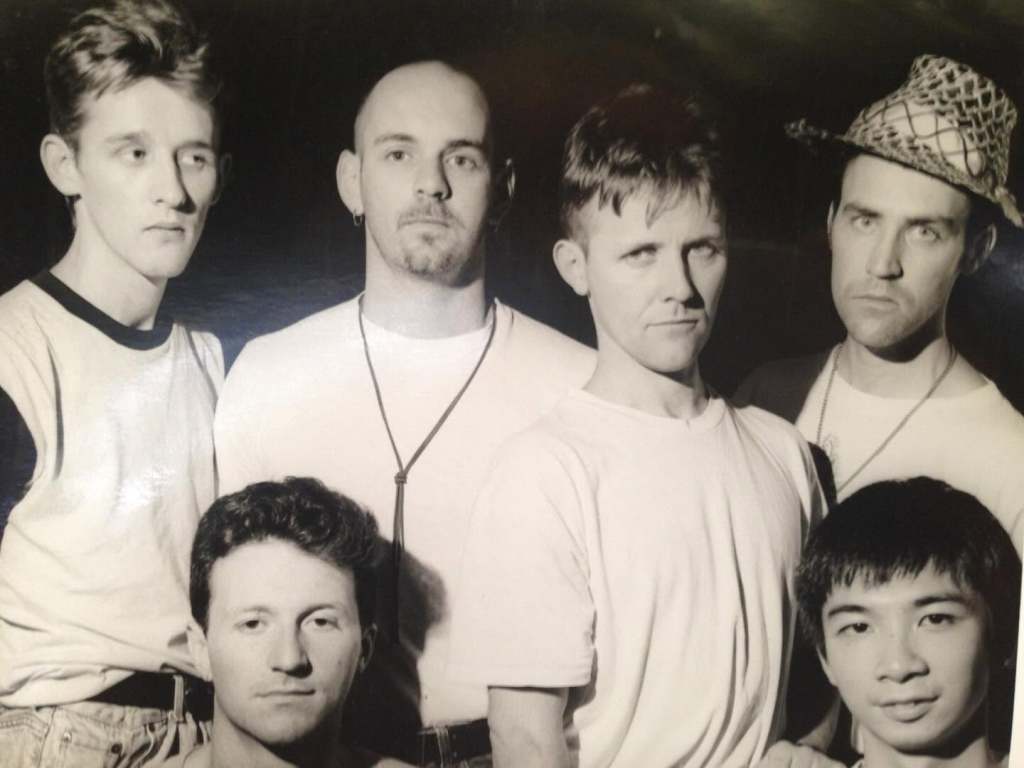 A black and white photo featuring six men posing together, wearing casual clothing. The group includes individuals with varied hairstyles and expressions, standing close to one another.