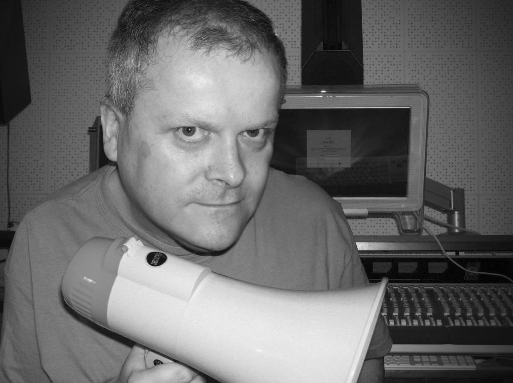 A man holding a megaphone in a studio setting, with a focused expression on his face. The image is in black and white.